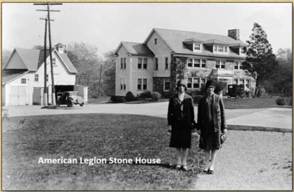 Historic view of the American Legion Stone House with two women standing on the lawn and early automobiles nearby